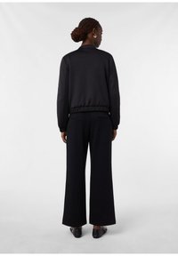Frau mit geflochtenem Haar, das zu einem Dutt gestylt ist, trägt eine schwarze Jacke, weite schwarze Hosen und schwarze Schuhe und steht vor einem weißen Hintergrund.