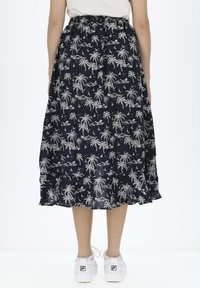 Marineblauwe maxi-rok met een palmbomen- en zeilbootprint, elastische tailleband, vloeiende stof en een geschulpte zoom. Gdragend met witte sneakers.