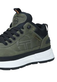 Grønn semsket sneaker med svarte mesh-detaljer, teksturerte svarte lisser, fremtredende logo på siden og hvit såle. Har en trekkflik.