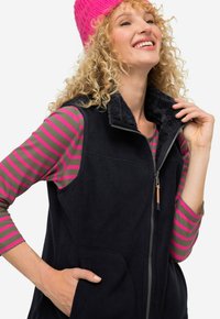 Czarny polarowy kamizelka z wysokim kołnierzem i zamkiem błyskawicznym, z bocznymi kieszeniami. Noszona na szarej i różowej, paskowanej koszuli z długim rękawem.
