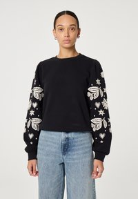 Zwarte sweatshirt met geborduurde witte bloemen- en sterrenpatronen op de mouwen, met een ronde halslijn en geribbelde manchetten. Gecombineerd met blauwe jeans.