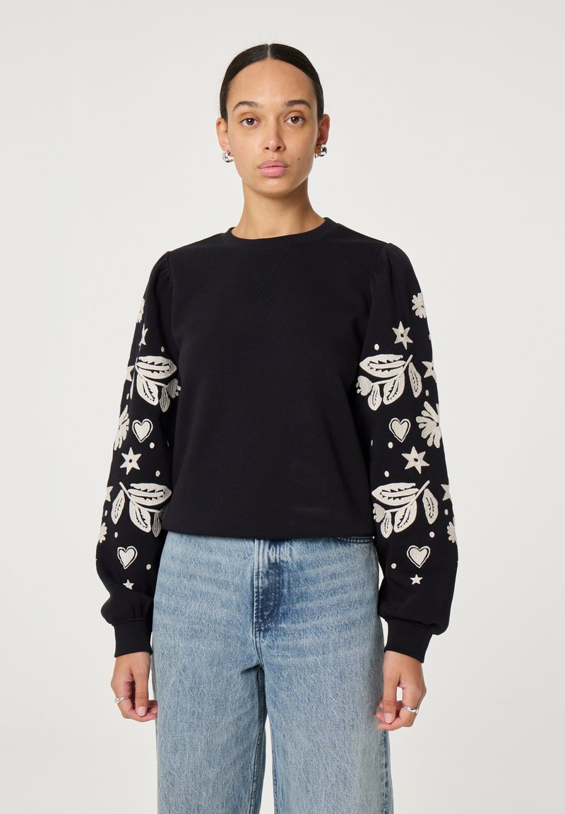 Zwarte sweatshirt met geborduurde witte bloemen- en sterrenpatronen op de mouwen, met een ronde halslijn en geribbelde manchetten. Gecombineerd met blauwe jeans.