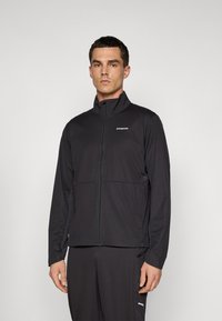 Man standing wearing a black Patagonia zip-up jacket and matching black pants against a plain light background.