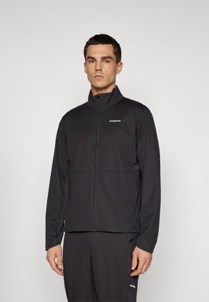 Man standing wearing a black Patagonia zip-up jacket and matching black pants against a plain light background.