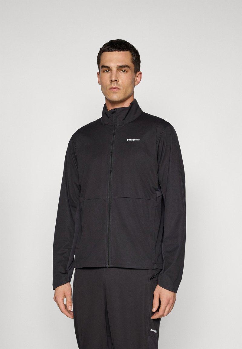 Man standing wearing a black Patagonia zip-up jacket and matching black pants against a plain light background.
