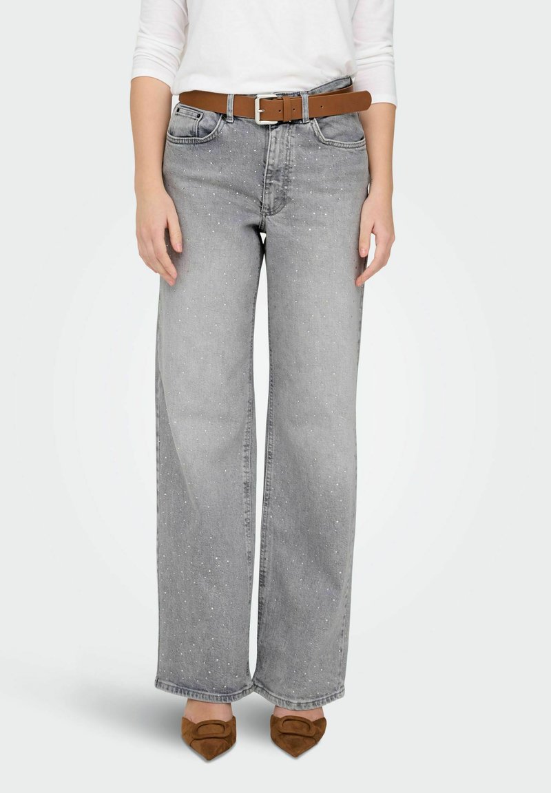 Jeans de mezclilla gris de pierna ancha con pequeñas manchas claras, combinados con un cinturón de cuero marrón con hebilla plateada y zapatos de tacón puntiagudos de gamuza marrón.