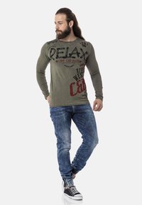Olivenfarbene Langarmshirt mit kräftigen schwarzen und roten Textdesigns, kombiniert mit blauen Jeans im Denim-Look mit ausgewaschenem Finish und schwarzen Sneakers.