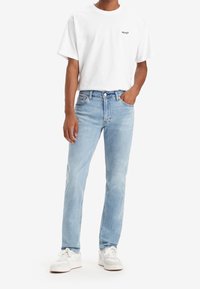 White short-sleeve t-shirt with Levi's logo, paired with light blue jeans. Model wears white sneakers with gray accents. Casual outfit.