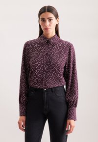 Bordeaux rood overhemd met witte stippen, een puntige kraag en lange mouwen. Draagt met hoge taille zwarte spijkerbroek.