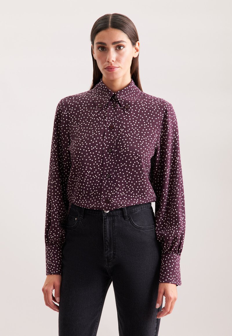 Bordeaux rood overhemd met witte stippen, een puntige kraag en lange mouwen. Draagt met hoge taille zwarte spijkerbroek.