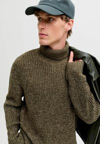 Maglione a collo alto in maglia marrone con un motivo strutturato, con dettagli a costine sul colletto e sui polsini. Il modello tiene una giacca di pelle nera.