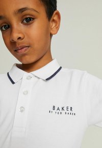 Biela polokošeľa s tmavo modrým lemovaním goliera a značkovým logom "BAKER BY TED BAKER" v tmavomodrej farbe. Obsahuje zapínanie na tri gombíky a textúrovanú látku.