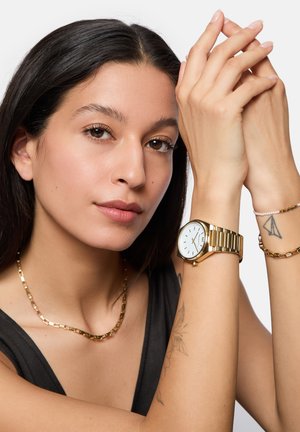 Goldene Metallarmbanduhr mit weißem Zifferblatt, getragen mit einer goldenen Kettenhalskette. Glatte Haut und sichtbare Tattoos am Unterarm.