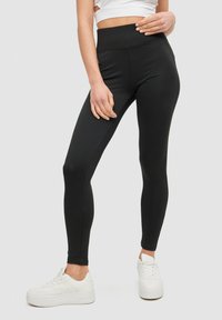 Mujer vestida con leggings negros de cintura alta y zapatillas de plataforma blancas, de pie contra un fondo liso.