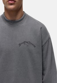 Felpa grigia con collo rotondo a costine, caratterizzata da un logo in testo nero sul davanti. Texture morbida, design casual.