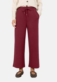 Burgunderfarbene, weit geschnittene Sweatpants mit elastischem Bund, Kordelzug, seitlichen Taschen und glatter Stofftextur. Kombiniert mit dunklen, mit Nieten verzierten Schuhen.