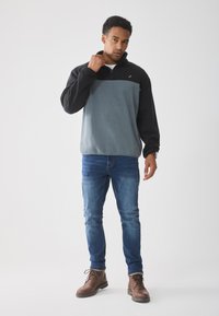 Sudadera de forro polar con media cremallera, que presenta una parte superior negra y una parte inferior gris claro. Lleva puesta con vaqueros azules y botas marrones con cordones.