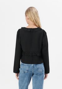 Black long-sleeve top with ruffled collar and hem, featuring a cropped design. Paired with light blue high-waisted jeans. Smooth texture.