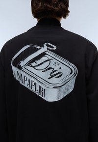 Fekete kapucnis pulóver, amelyen egy fémdoboz grafikája látható, rajta a "Drip" és "NAPAPIJRI" felirat szürkében. A dizájn magában foglal egy kihúzó fület is.