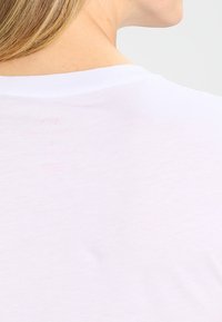 Bílá tričko s hladkou texturou a kulatým výstřihem. Na zadní straně, poblíž oblasti krku, se nachází decentní růžový potisk.