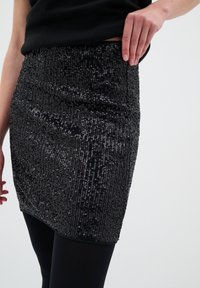 Mini jupe noire en sequins avec une coupe ajustée, présentant une surface texturée et une taille lisse, associée à des leggings noirs.
