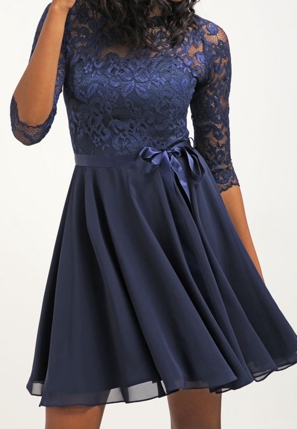 Femme portant une robe bleu marine longueur genou avec un corsage en dentelle, des manches trois-quarts et un ruban en satin noué à la taille.