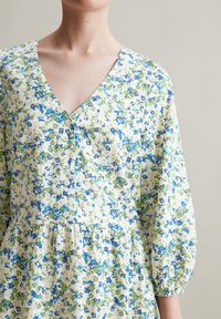 Blumenkleid mit V-Ausschnitt, das blau-grüne Muster auf einem hellen Hintergrund zeigt, mit Knopfdetails und Puffärmeln. Weicher Stoff.