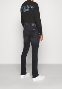 Camisa negra de manga larga con texto azul en la parte posterior, combinada con jeans oscuros ajustados y zapatillas blancas, mostrando un atuendo casual.