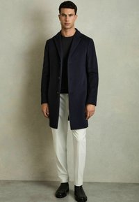 Mørkeblå uldfrakke med enkelt brystdesign, opretstående revers og tre knapper, parret med hvide bukser og sorte ankelstøvler.