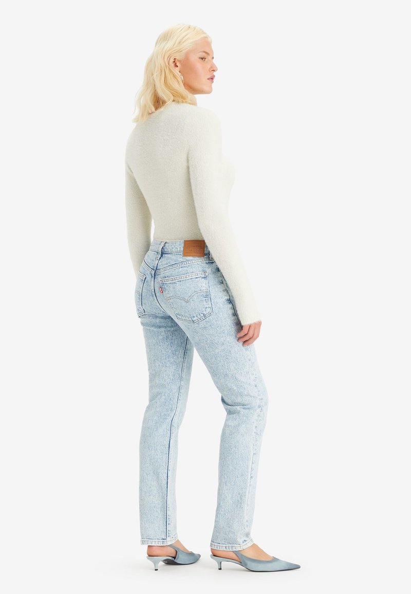 Pull en laine gris clair et flou, jean taille haute bleu clair avec une patch en cuir beige, et escarpins bleus à bout pointu. Le modèle fait dos à l'appareil photo.