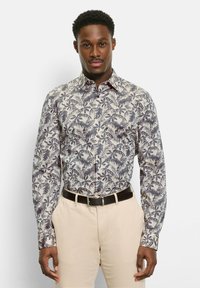 Mann trägt ein beiges Hemd mit dunklen Blattmustern, beige Hose und schwarzen Gürtel, steht vor einem schlichten weißen Hintergrund.