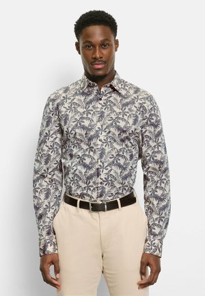 Mann trägt ein beiges Hemd mit dunklen Blattmustern, beige Hose und schwarzen Gürtel, steht vor einem schlichten weißen Hintergrund.