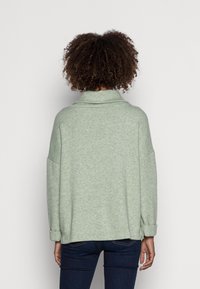 Pull vert clair oversize avec col plié et épaules tombantes ; fabriqué en tissu doux et texturé ; poignets visibles retroussés aux manches.
