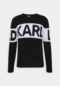Musta neulepaita, jossa on rohkea valkoinen raita ja teksti "KARL". Pyöreä pääntie ja pitkät hihat, joissa resorit hihansuissa ja helmassa.