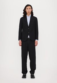 Schwarzer maßgeschneiderter Anzug mit einem einreihigen Blazer und weiten Hosen. Darunter ein hellblaues Hemd, kombiniert mit schwarzen Schuhen.