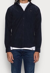 Camicia a maniche lunghe color blu navy, in misto cotone, con chiusura frontale a bottoni, tessuto morbido, vestibilità slim e logo discreto sul petto, abbinata a una maglietta bianca sotto.