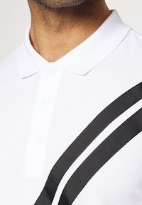 Bílá polo košile vyrobená z bavlny, s klasickým límečkem, třemi knoflíky a dvěma diagonálními černými pruhy přes hrudník.
