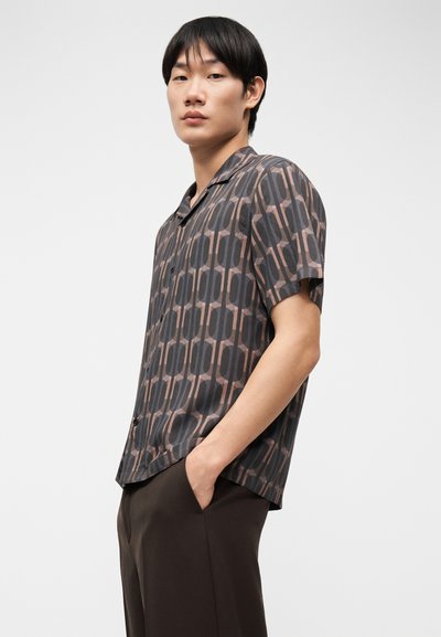 Νέος άνδρας με κοντά μαύρα μαλλιά, ντυμένος με πουκάμισο κοντομάνικο με σχέδιο και σκουρόχρωμο παντελόνι, στέκεται με το ένα χέρι στην τσέπη.