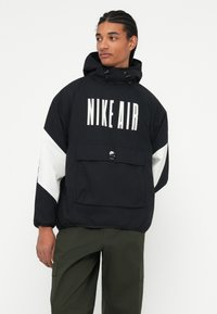 Juodas vėjui atsparus paltas su baltais akcentais, su priekinia kišene, reguliuojamu kapu ir ryškiai matomu „NIKE AIR“ logotipu priekyje.