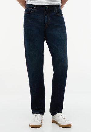 Person iført mørkeblå jeans med lige ben, hvide sneakers og en stribet skjorte, stående mod en hvid baggrund.