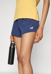 Άτομο που φορά μπλε σορτς New Balance και κίτρινο μπλουζάκι, κρατώντας ένα μαύρο μπουκάλι νερού στο πλάι του, μπροστά από απλό φόντο.