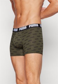 Olivgrüne Boxershorts mit enger Passform, ausgestattet mit einem schwarzen elastischen Bund, der mit weißem "PUMA"-Branding versehen ist. Auffälliges, sich wiederholendes Logo-Muster.
