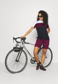 Tenue de cyclisme avec un schéma de couleurs bleu marine et bordeaux ; haut à manches courtes avec une fermeture éclair sur le devant, associé à un short assorti. Vélo de route argent.