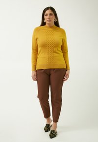 Maglione di maglia senape con pannello centrale testurizzato, abbinato a pantaloni aderenti marroni e scarpe nere con punta a stiletto decorate con borchie dorate.