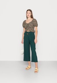 Blusa de flores escuras com mangas bufantes, combinada com calças largas verdes de cintura alta, sandálias amarelas e uma bolsa de ombro branca.