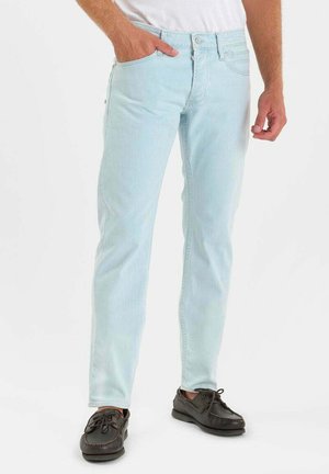 Mann trägt hellblaue Slim-Jeans mit schwarzen Schnürschuhen, eine Hand in der Tasche, steht vor einem weißen Hintergrund.