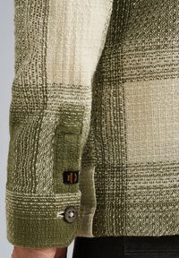 Close-up van een groene en beige geweven stoffen mouw met een zwarte knoop en een klein zwart PME Legend merkteken.