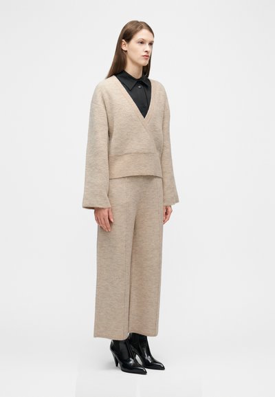 Γυναίκα που φοράει μπεζ πλεκτό παντελόνι φαρδιάς γραμμής και ταιριαστό πλεκτό πουλόβερ με V λαιμόκοψη πάνω από μαύρο πουκάμισο με γιακά, με μαύρες μποτάκια στον αστράγαλο.