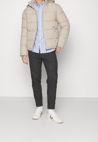 Beżowa kurtka puchowa z poziomym pikowaniem, z kapturem i kieszeniami na zamek, połączona z jasnoniebieską koszulą w paski i ciemnymi spodniami. Białe trampki.