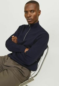 Pulover tricotat bleumarin cu guler înalt și fermoar argintiu; modelul așezat pe un scaun metalic, cu brațele încrucișate, purtând pantaloni bej deschis.